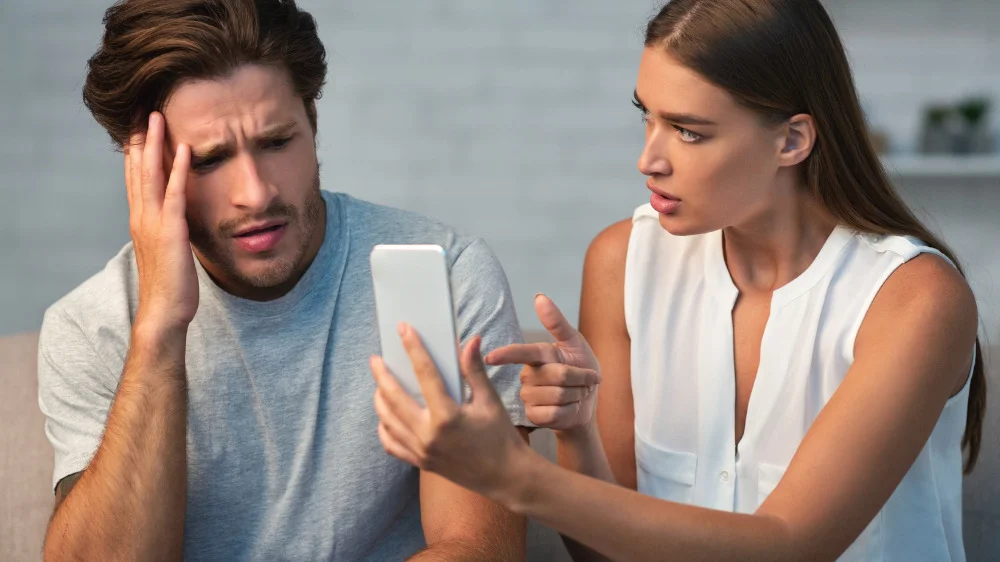 右手で男性の白いスマートフォンを指差しながら怒っている女性