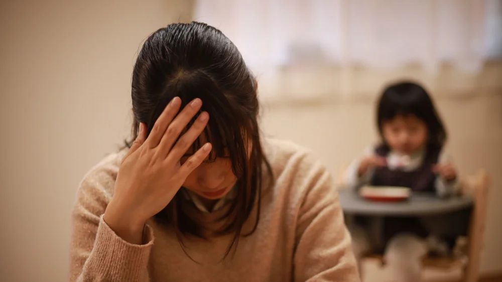 右手でおでこを抑えながら下を向く女性と後ろの子供