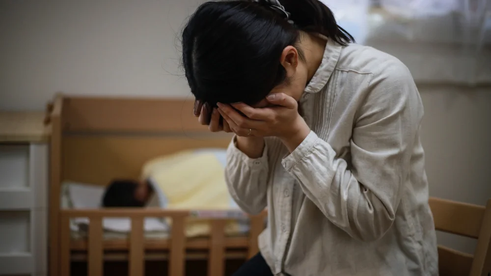 両手で顔を覆いながら俯く女性と後ろで寝ている赤ちゃん